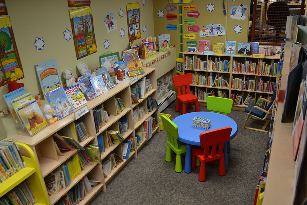 À l&apos;intérieur de la bibliothèque Champlain, un aperçu de la zone des enfants avec une bibliothèque de livres pour enfants et un petit coin salon avec table et chaise